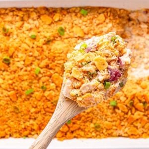 A wooden spoon scoops cheesy Ritz Cracker Chicken Casserole with a golden cracker topping from a white baking dish.