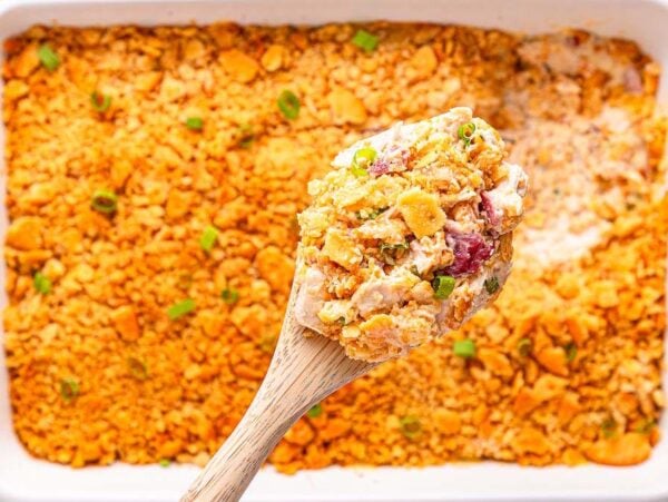 A wooden spoon scoops cheesy Ritz Cracker Chicken Casserole with a golden cracker topping from a white baking dish.