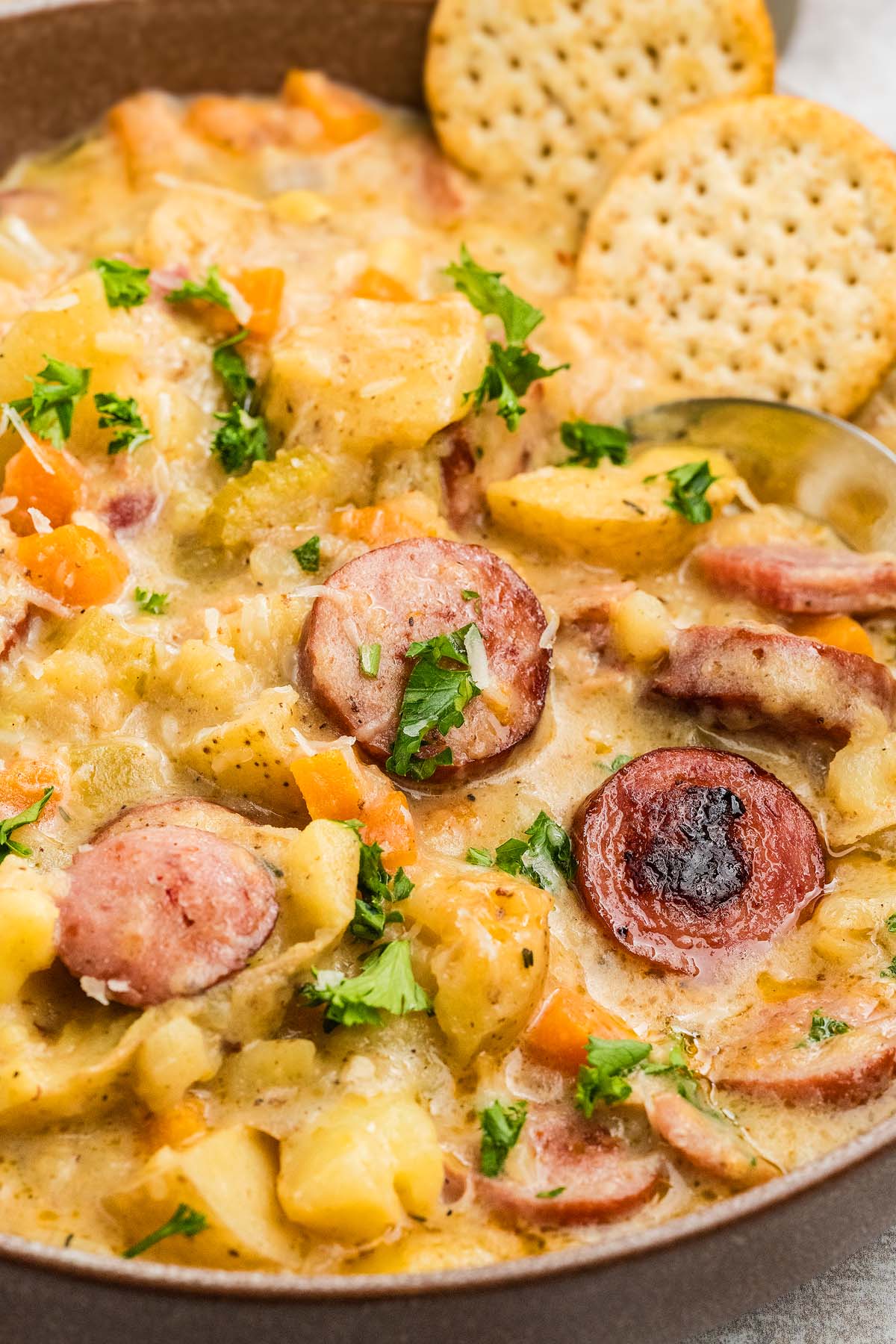 A creamy sausage potato soup with sliced sausage, potatoes, carrots, herbs, and two round crackers on the side.
