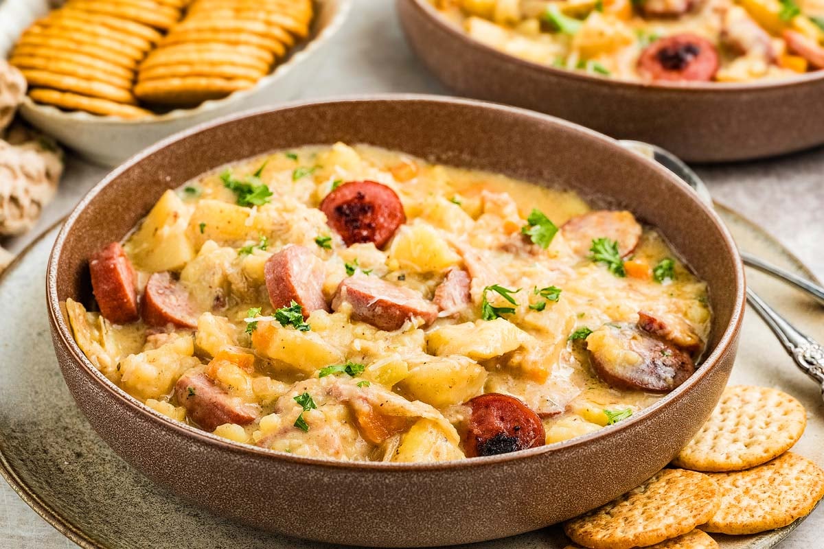 A bowl of hearty sausage potato soup garnished with herbs, served with round crackers on the side.