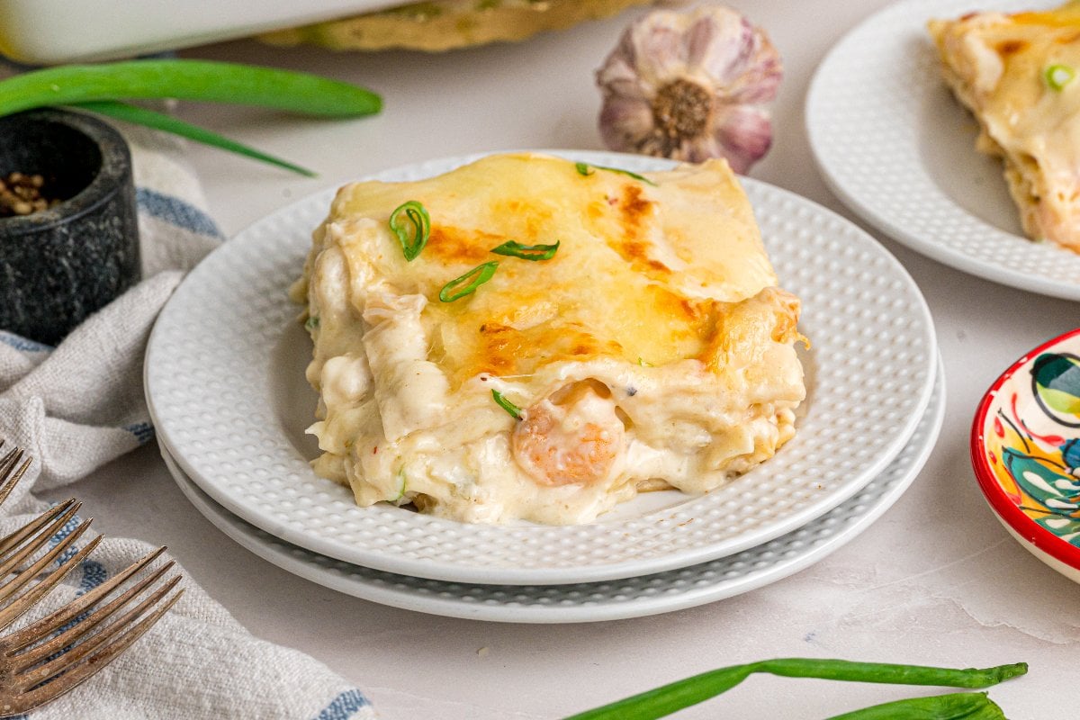 A slice of creamy seafood lasagna, garnished with green herbs, sits elegantly on a white plate.