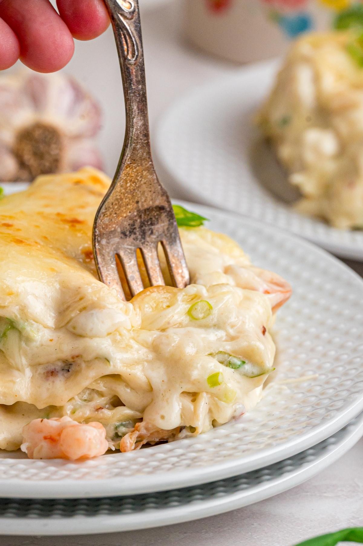 A fork cutting into creamy seafood lasagna on a white plate.