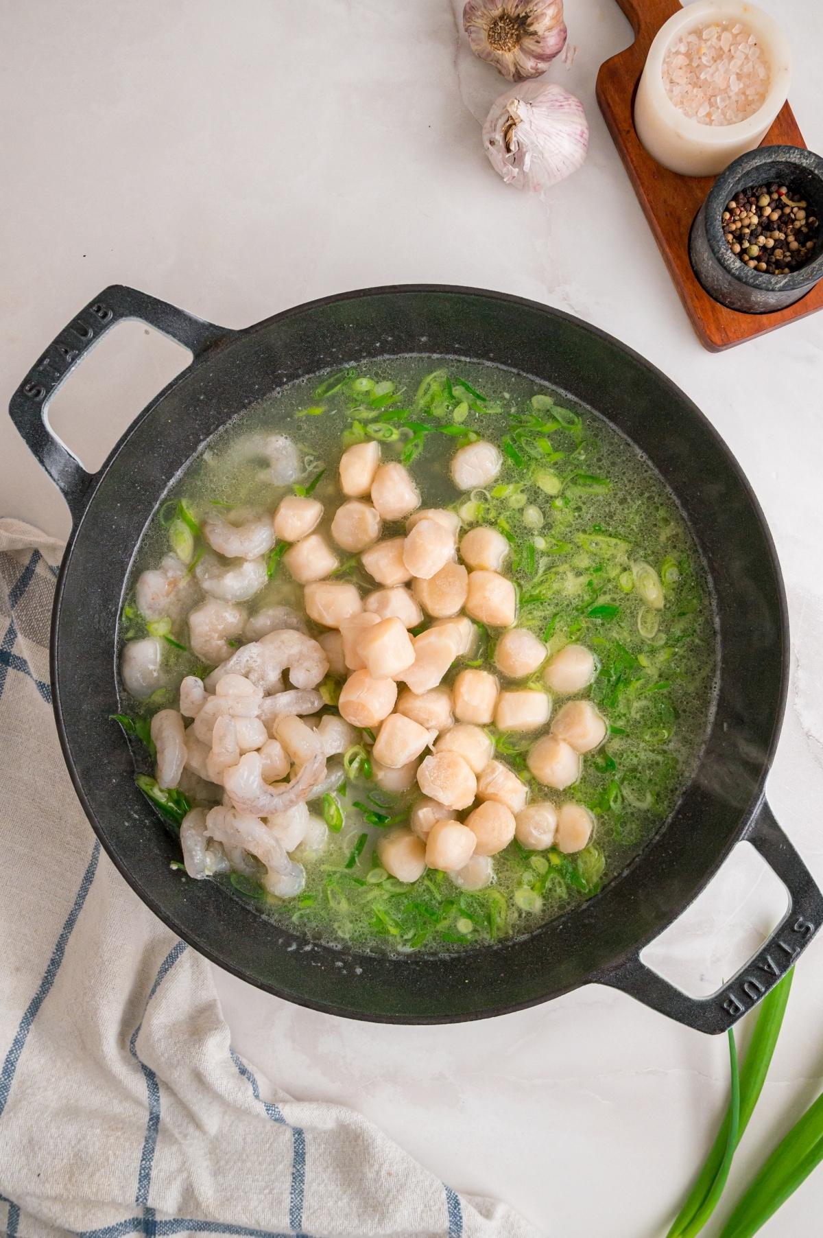 A black pot with shrimp, scallops, and green onions cooking in broth, next to garlic and spices.