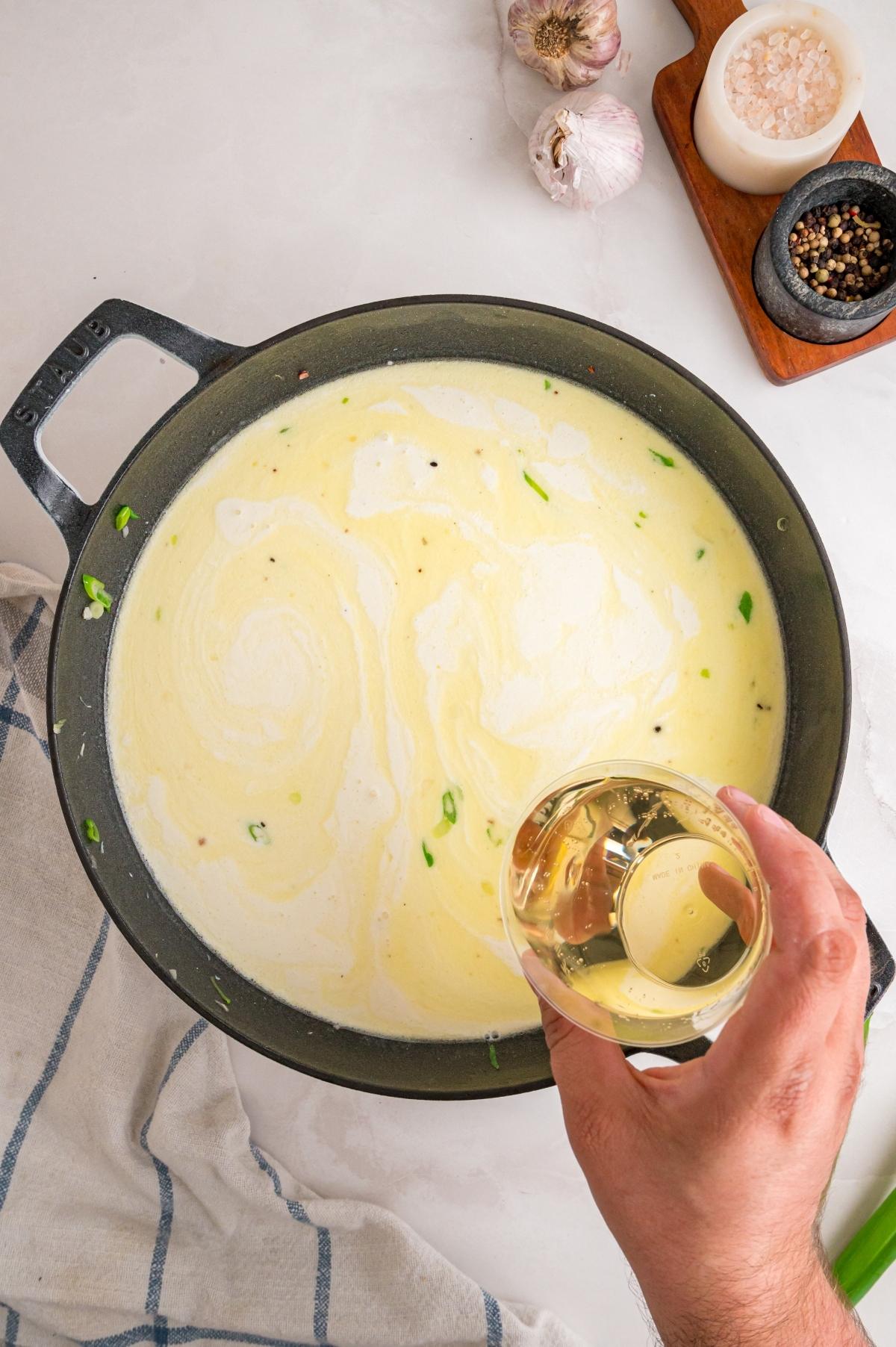 A hand pours liquid into a creamy sauce in a black pan, surrounded by garlic, salt, and pepper.