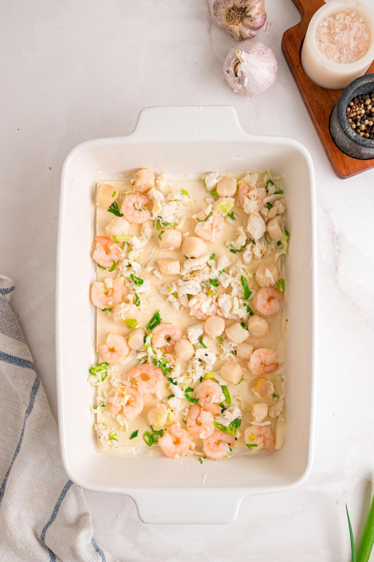 Rectangular baking dish with raw shrimp, scallops, and crab in a creamy white sauce, ready to bake.