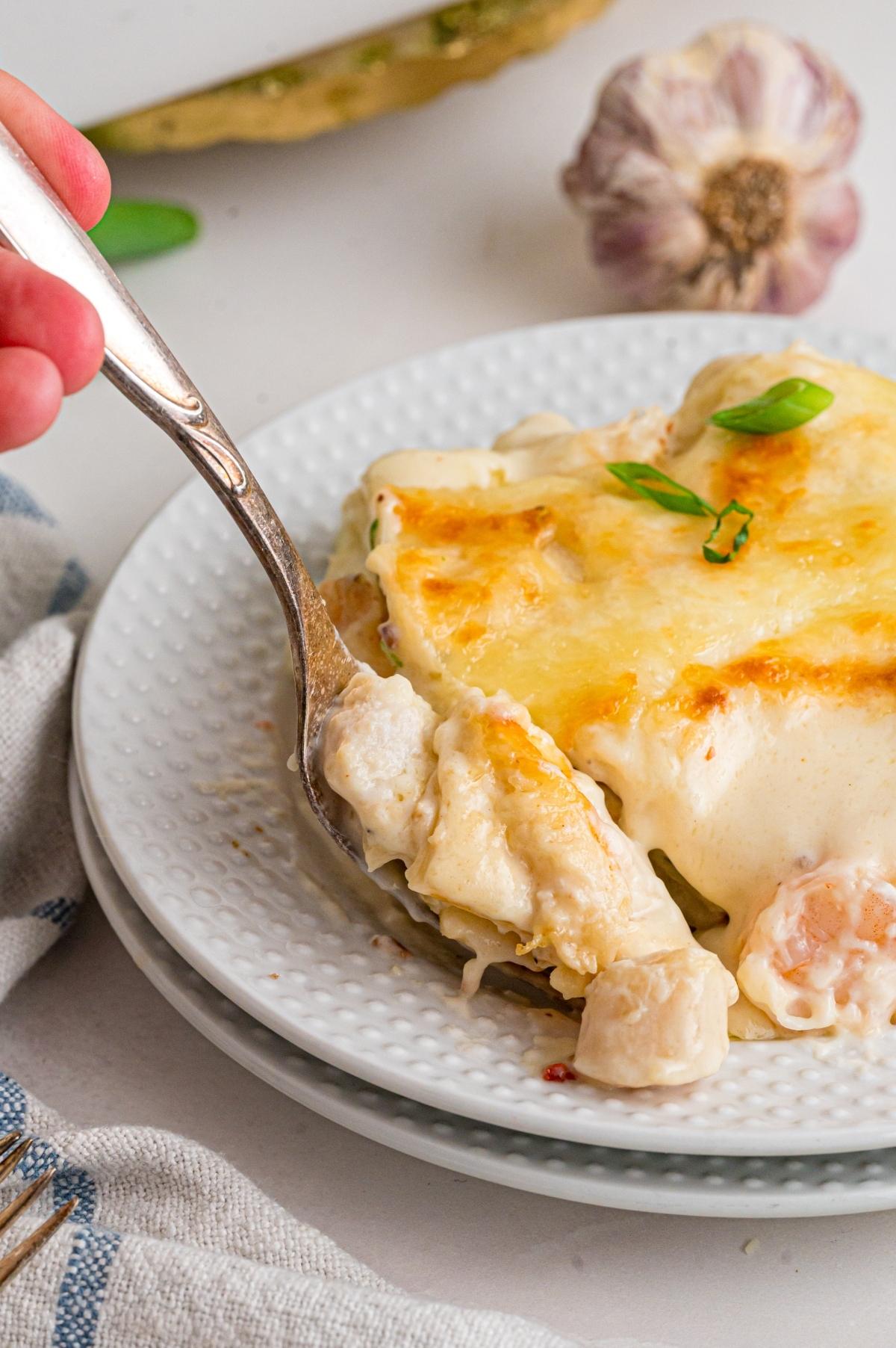 A fork scooping creamy seafood lasagna from a white plate, garnished with green herbs.