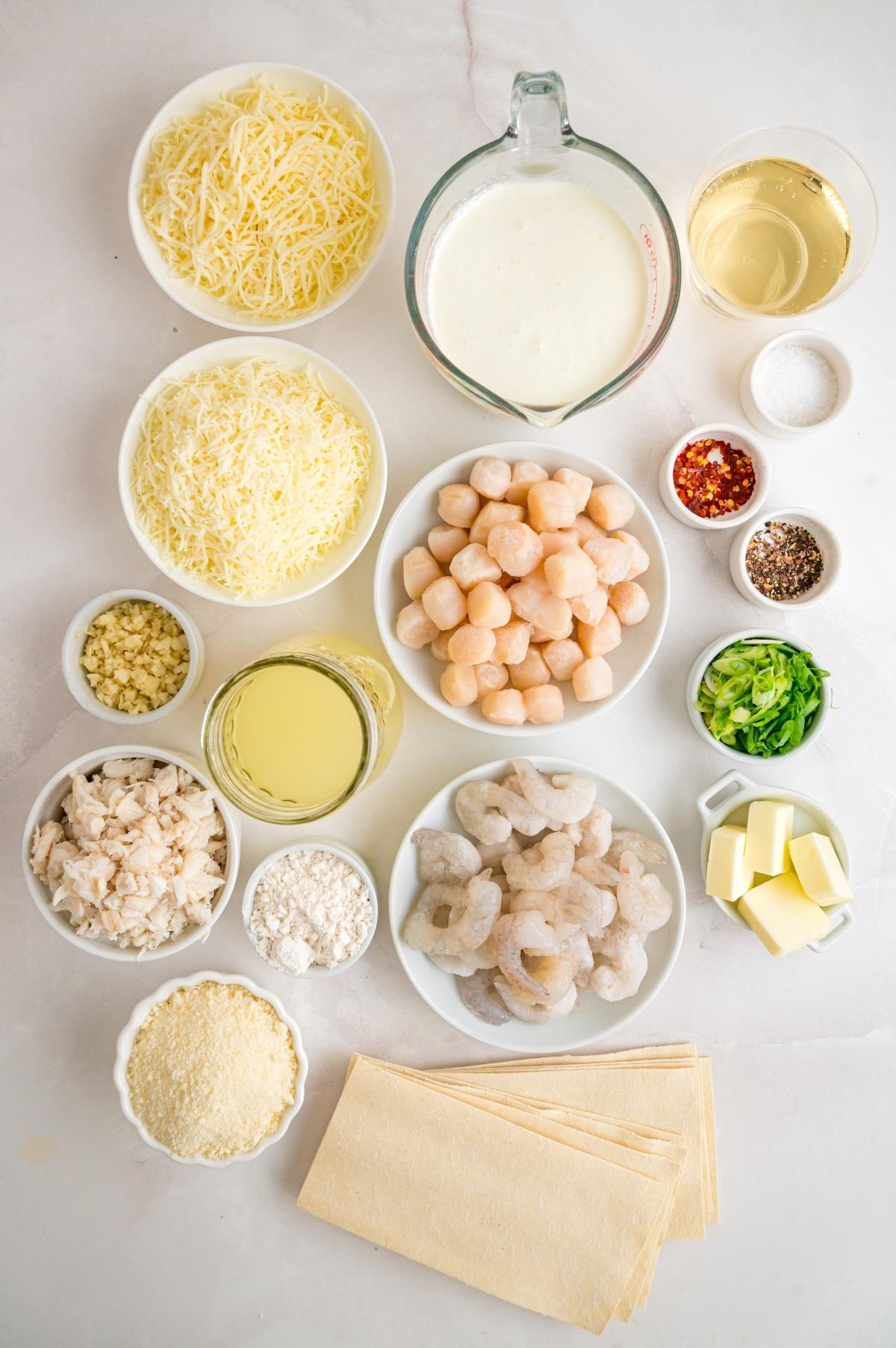 Various bowls hold cheese, cream, scallops, shrimp, spices, butter, lasagna sheets, and other ingredients on a counter.