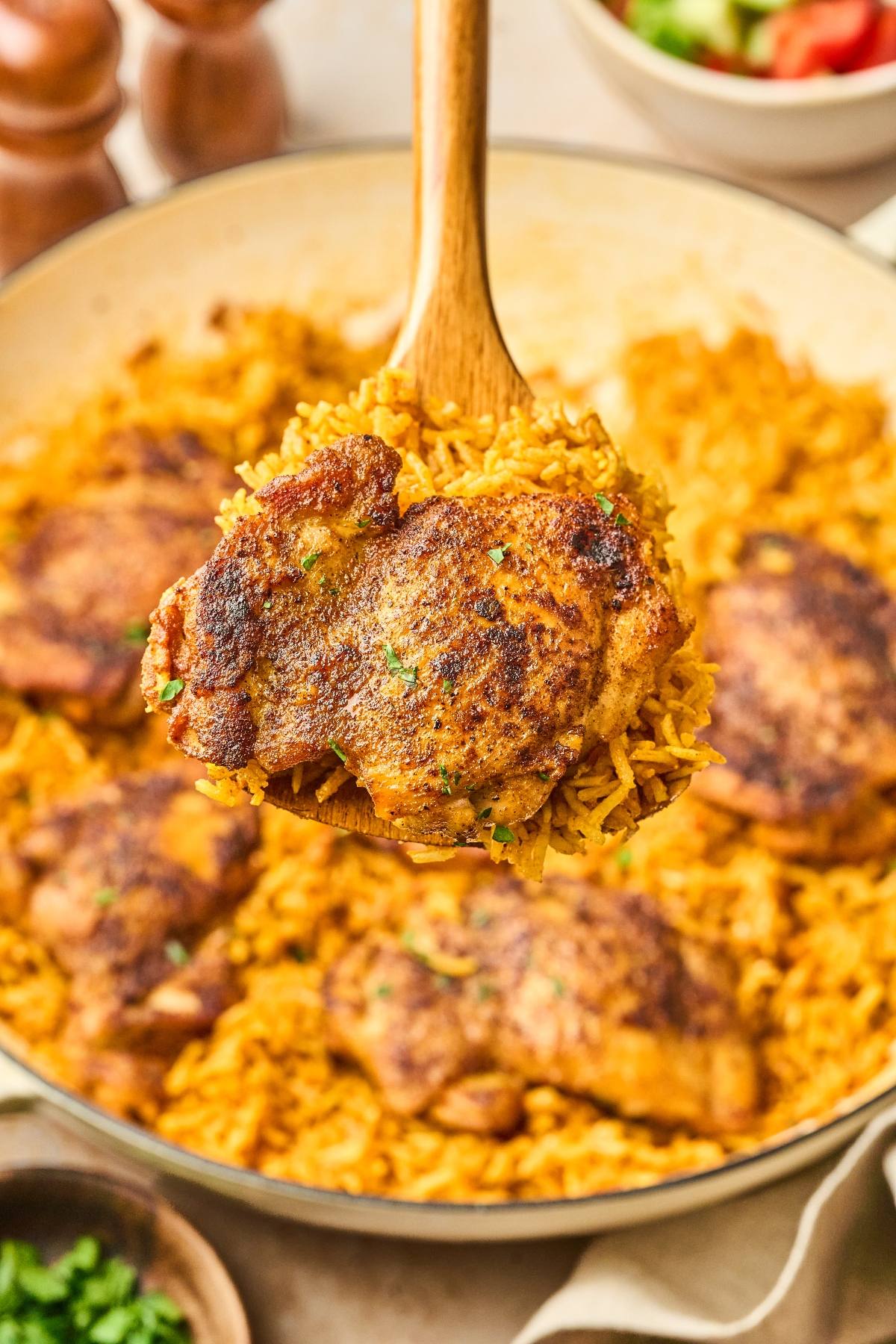 A wooden spoon holds seasoned chicken thigh over a pan of yellow rice and more chicken thighs.