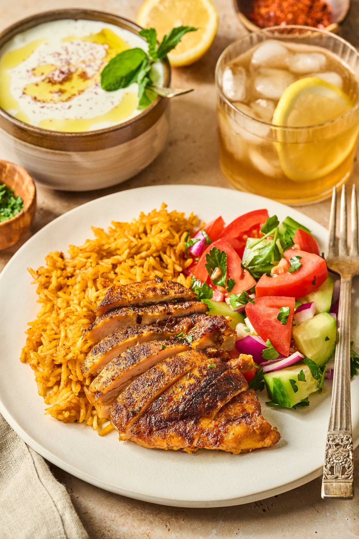 A plate with sliced grilled chicken, spiced rice, and a cucumber-tomato salad, with iced tea and dip on the side.