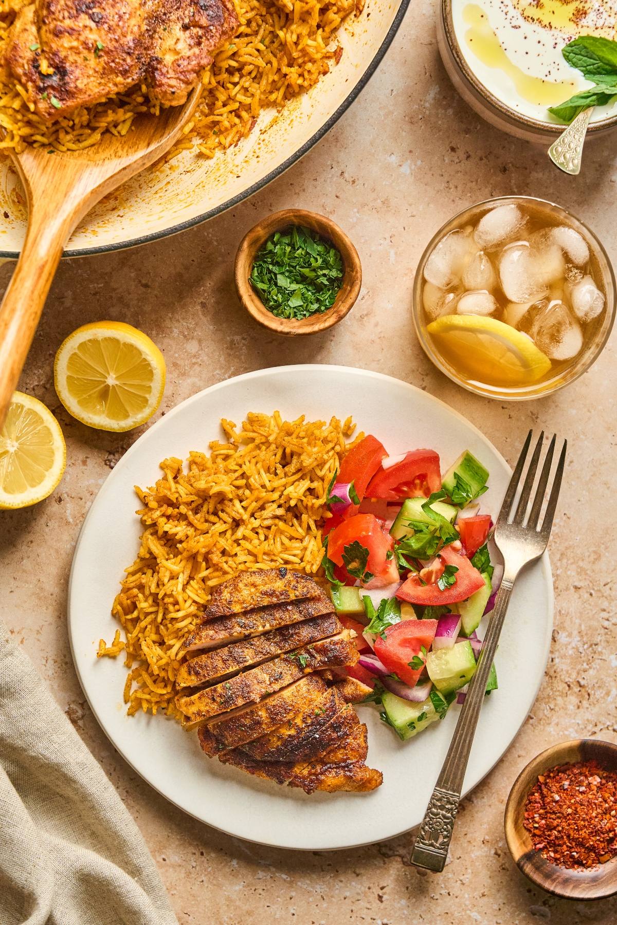 A plate with spiced rice, grilled chicken, salad, and a fork, with iced tea, lemon, and herbs nearby.