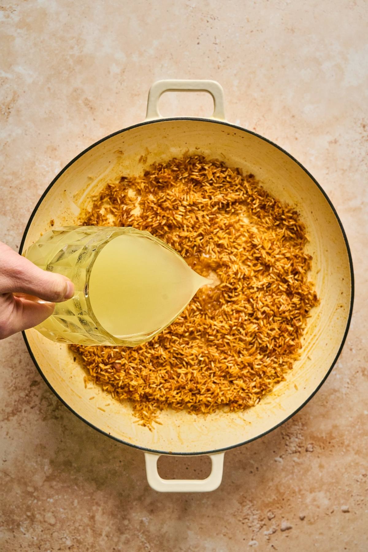 A hand pours broth from a measuring cup into a pot of toasted rice on a beige surface.