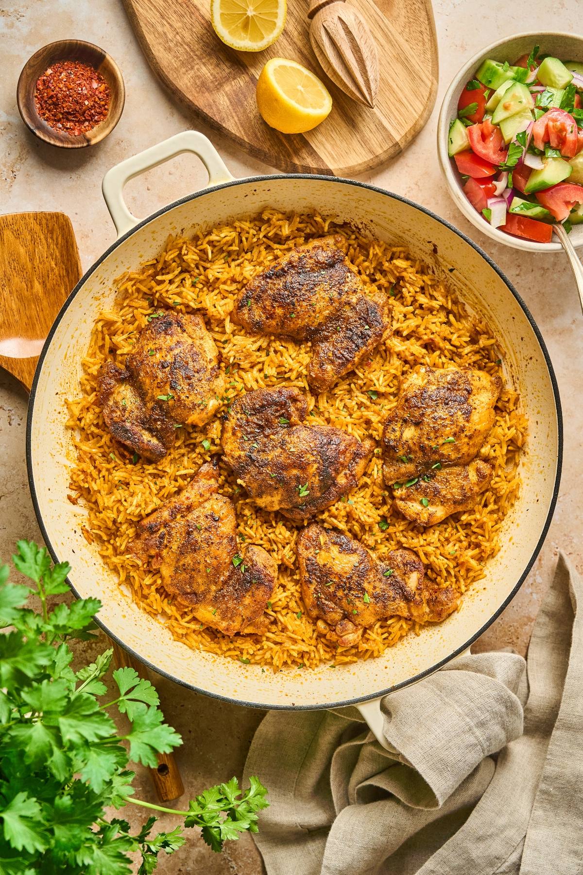 A pan of seasoned chicken thighs on yellow rice, with salad, lemon, and spices on the side.