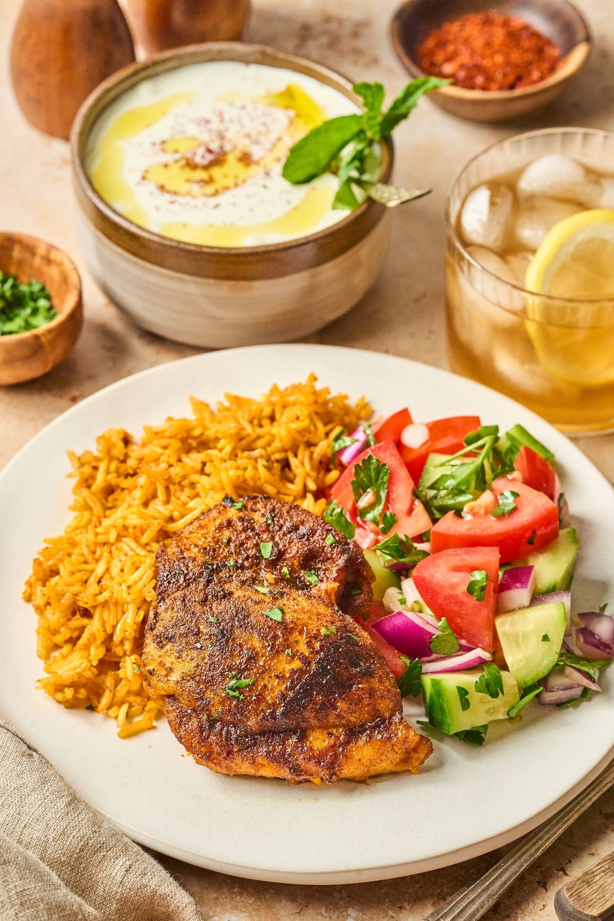 Plate with spiced chicken, yellow rice, cucumber-tomato salad; yogurt dip and iced tea in the background.