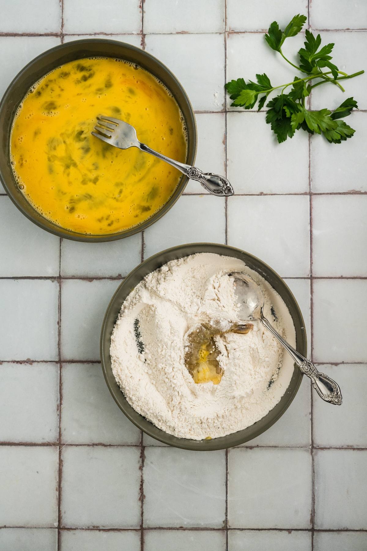 Two bowls with beaten eggs and flour mixture, each with a fork; parsley on the side on a tiled surface.