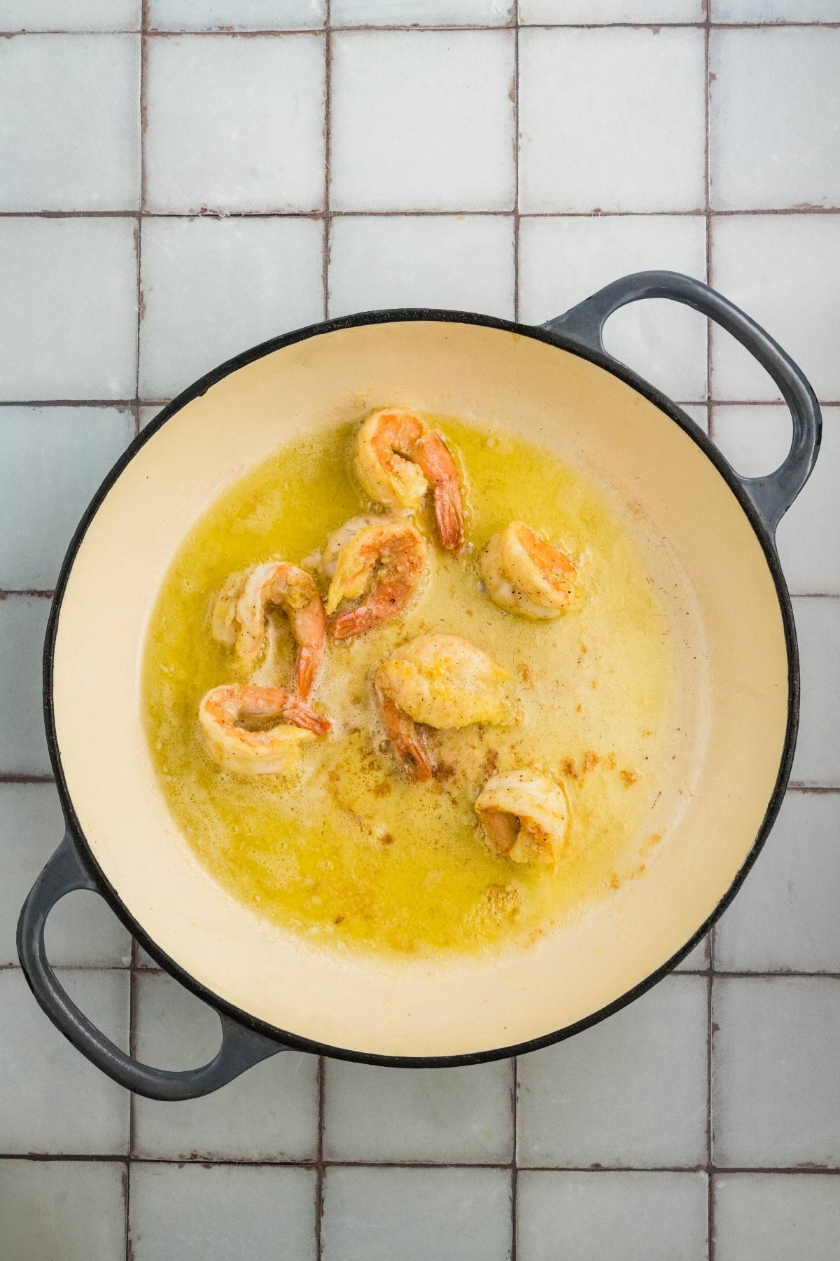 Shrimp cooking in a creamy garlic butter sauce in a round, white skillet on a tiled surface.