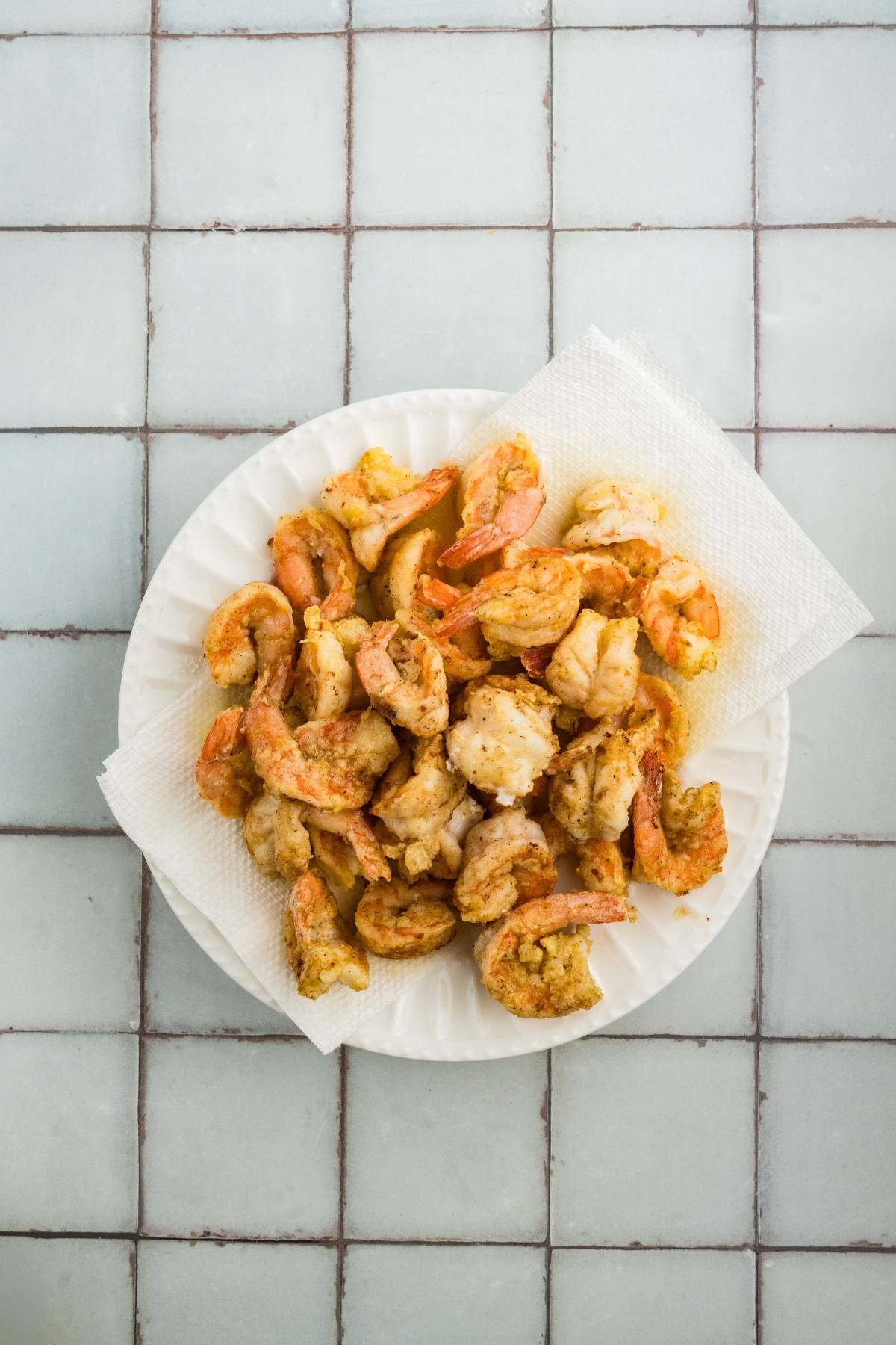 A white plate with fried shrimp on a paper towel, set on a tiled surface.