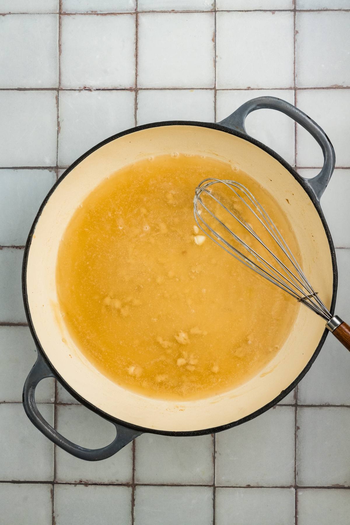 A whisk in a pot with melted butter on a white tiled countertop.