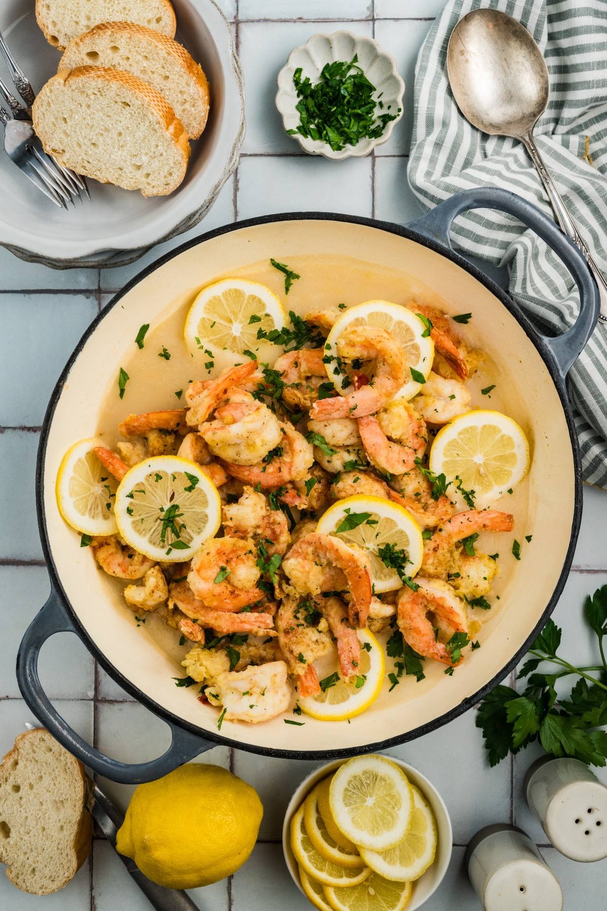 A pan of shrimp garnished with lemon slices and parsley, surrounded by bread, lemon, and herbs.