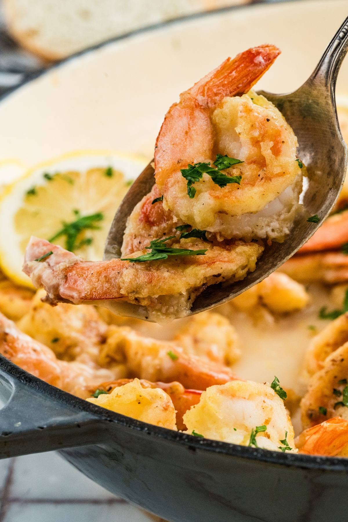 A spoon lifts breaded shrimp garnished with parsley from a pan, with lemon slices in the background.