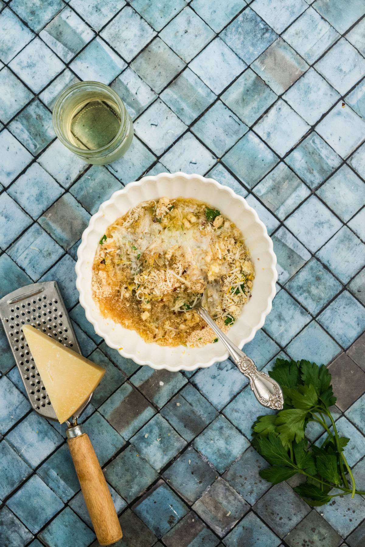 Overhead view of a bowl of risotto with a fork, cheese wedge, grater, parsley, and a glass of white wine.