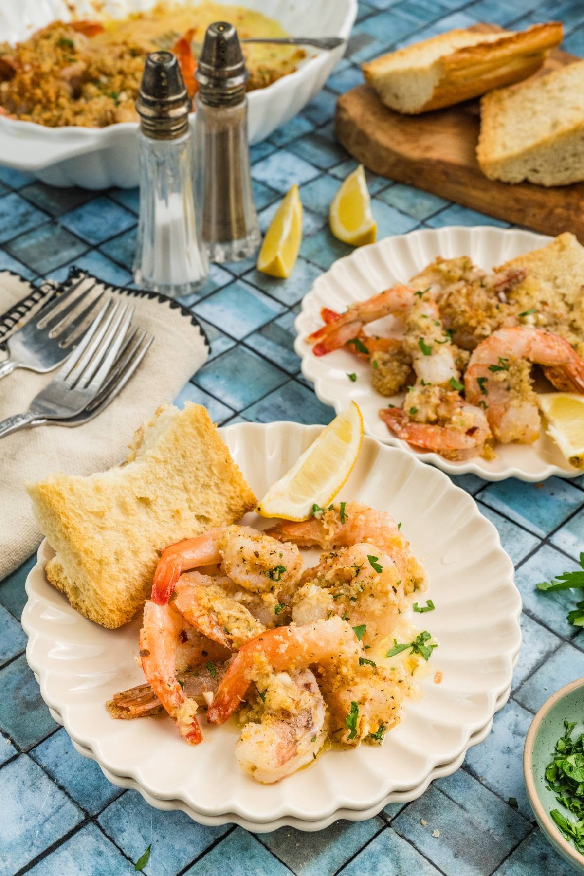 Plates of baked shrimp with breadcrumbs, lemon wedges, and bread slices on a blue tile table.