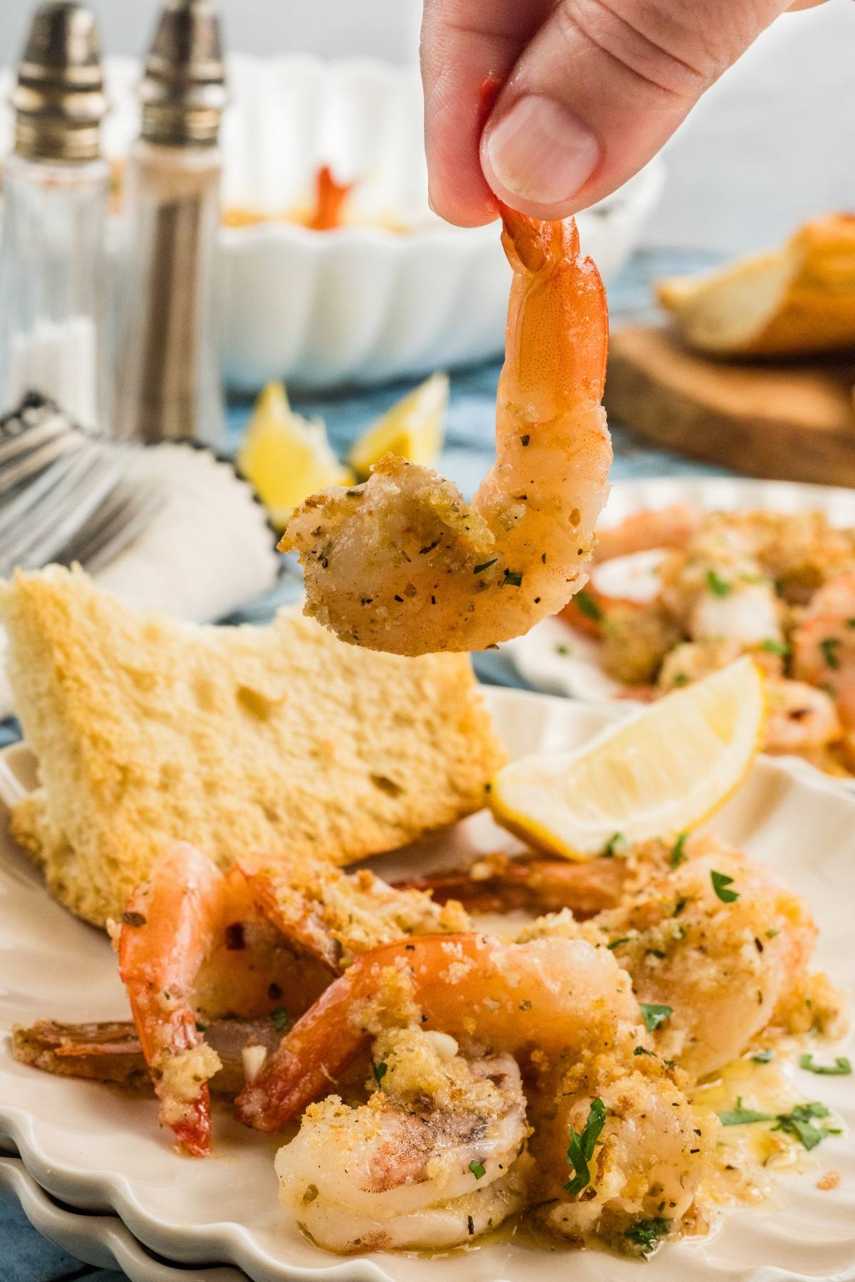 A hand holds a seasoned shrimp above a plate with shrimp, bread, lemon wedge, and herbs.