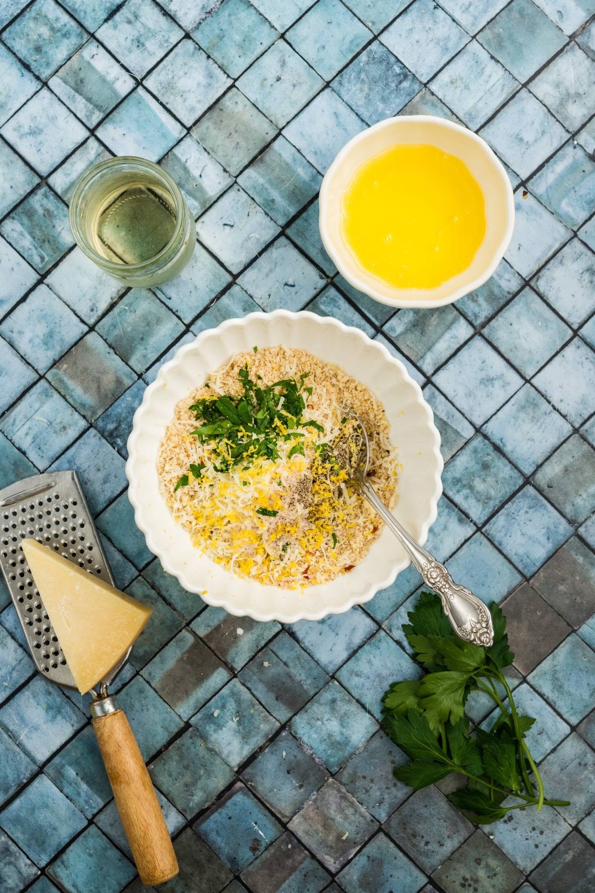 Overhead view of grated cheese, a bowl of breadcrumbs with herbs, melted butter, and a glass of oil on blue tiles.