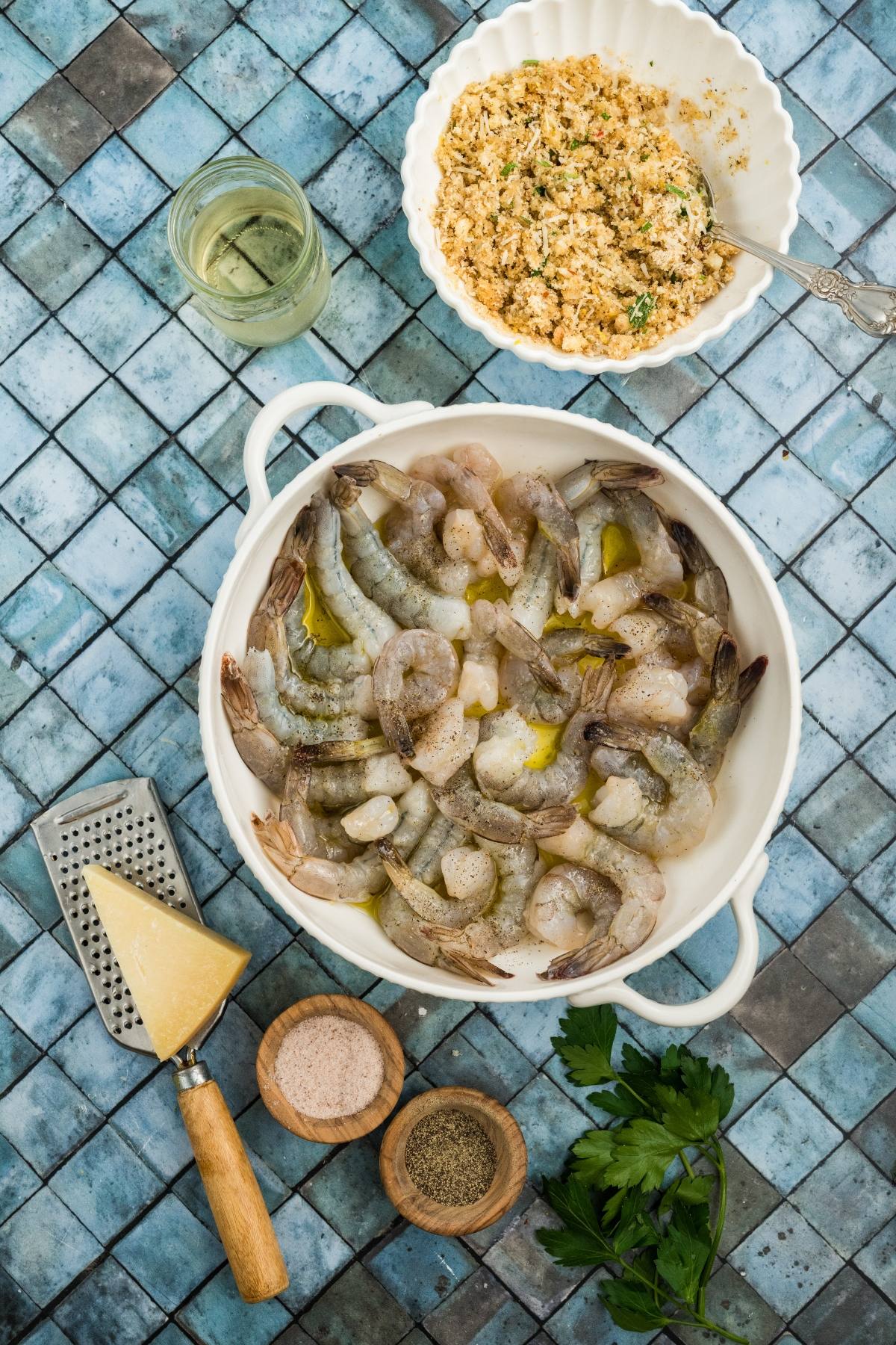 Raw shrimp in a white dish with seasonings, cheese, herbs, and a bowl of breadcrumbs on a tiled surface.