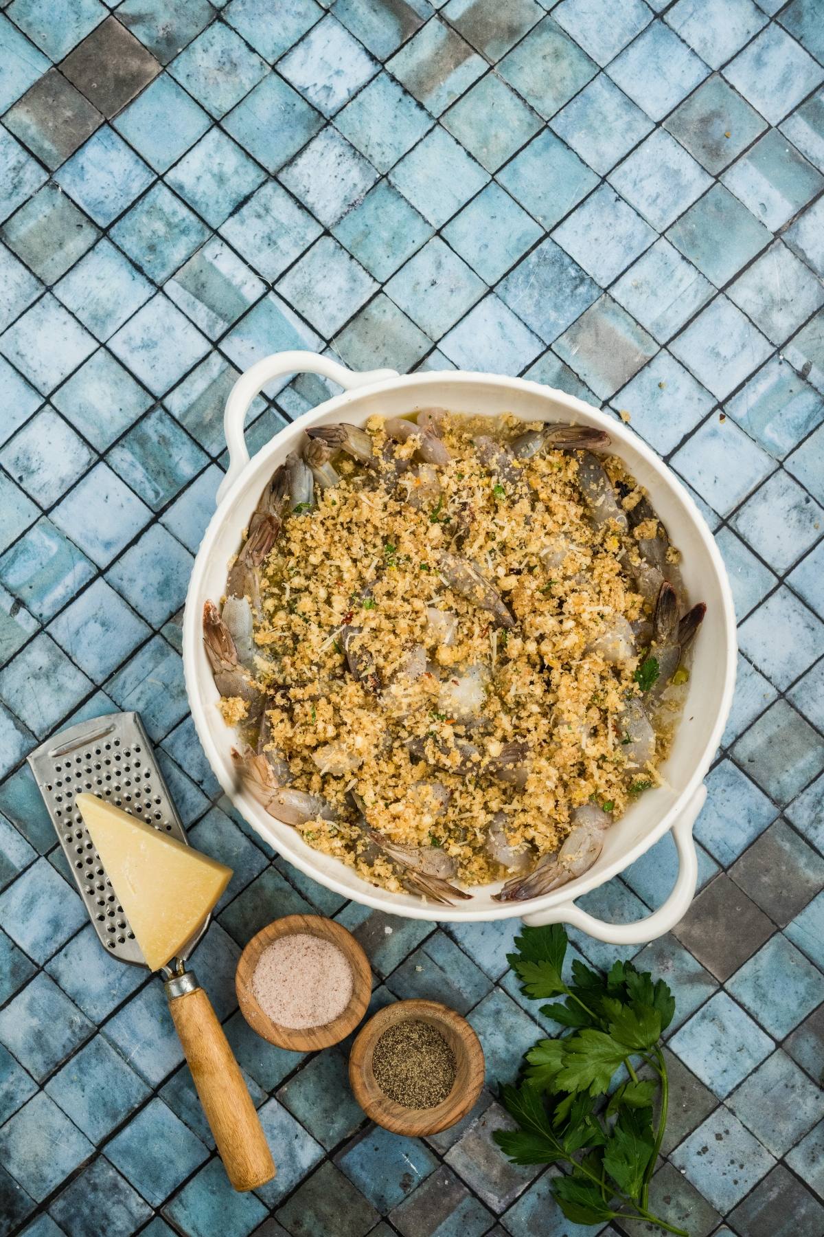 White dish of shrimp topped with garlic crumbs, with cheese, salt, pepper, and parsley on blue tile background.
