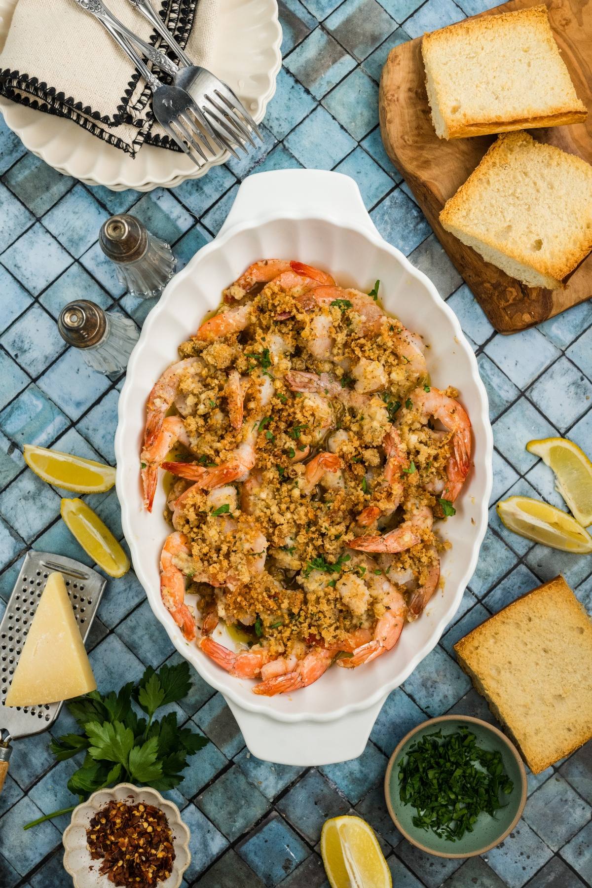 Baked shrimp with breadcrumbs in a white dish, surrounded by lemon wedges, toast, and herbs on a blue tile table.