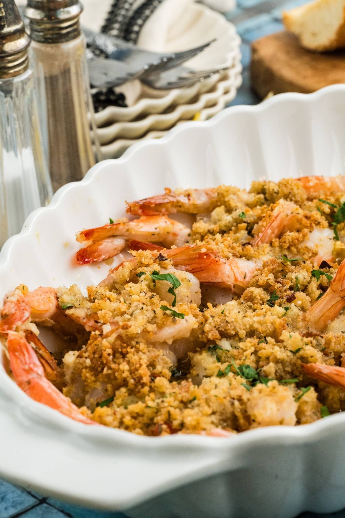 Baked shrimp with golden breadcrumb topping in a white dish, served with plates and utensils in the background.