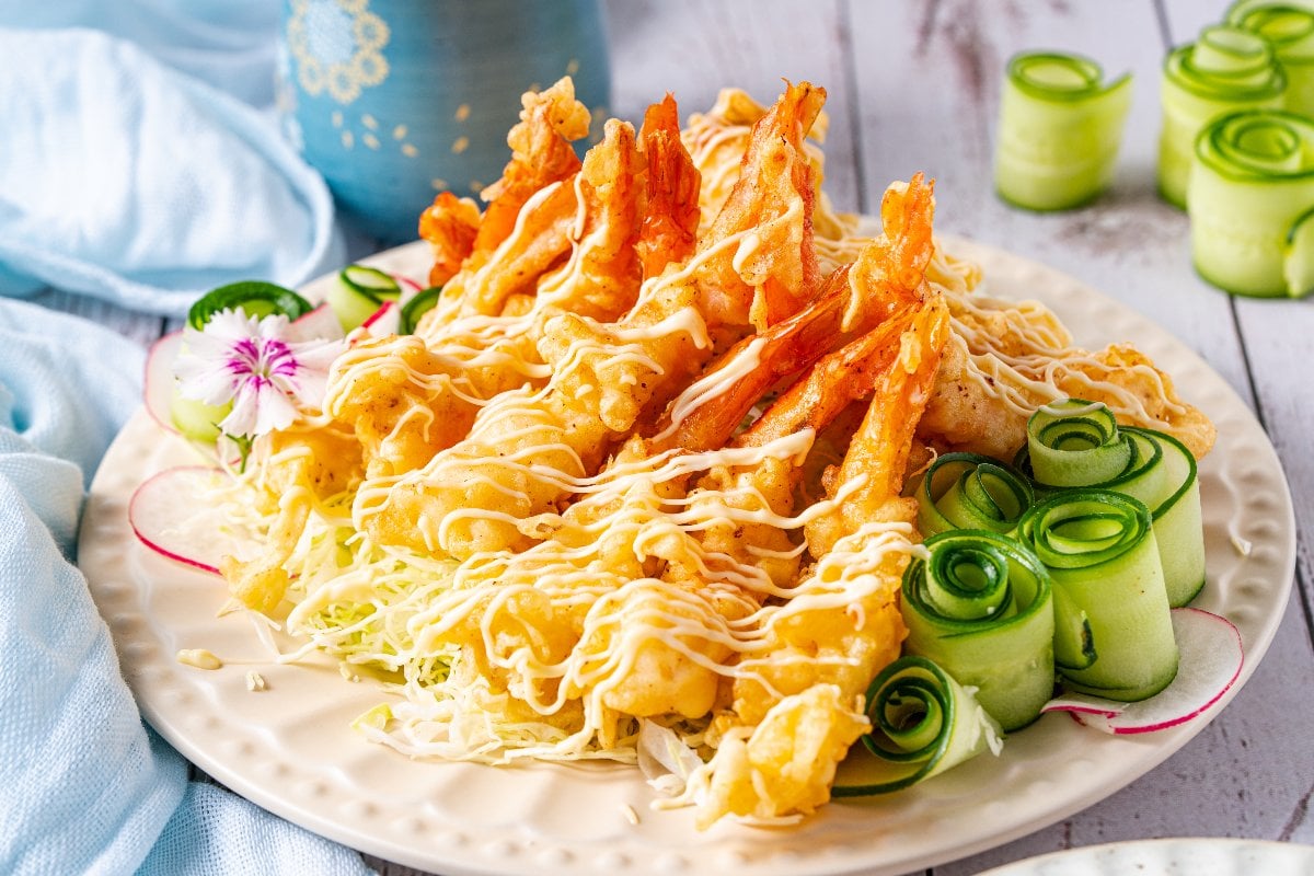 Plate of crispy shrimp tempura drizzled with sauce, served with cucumber rolls and garnished with a delicate flower.