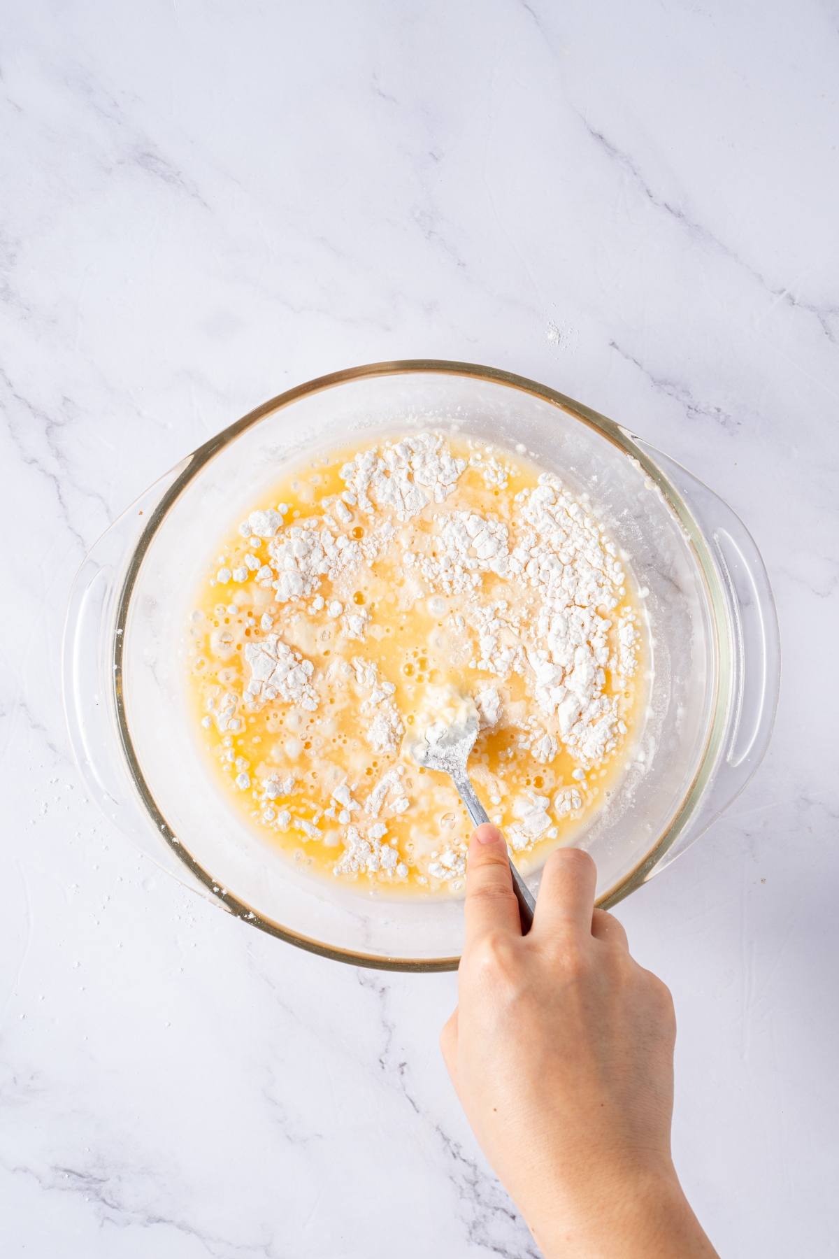 A hand mixing flour, eggs, and liquid in a glass bowl on a marble surface.