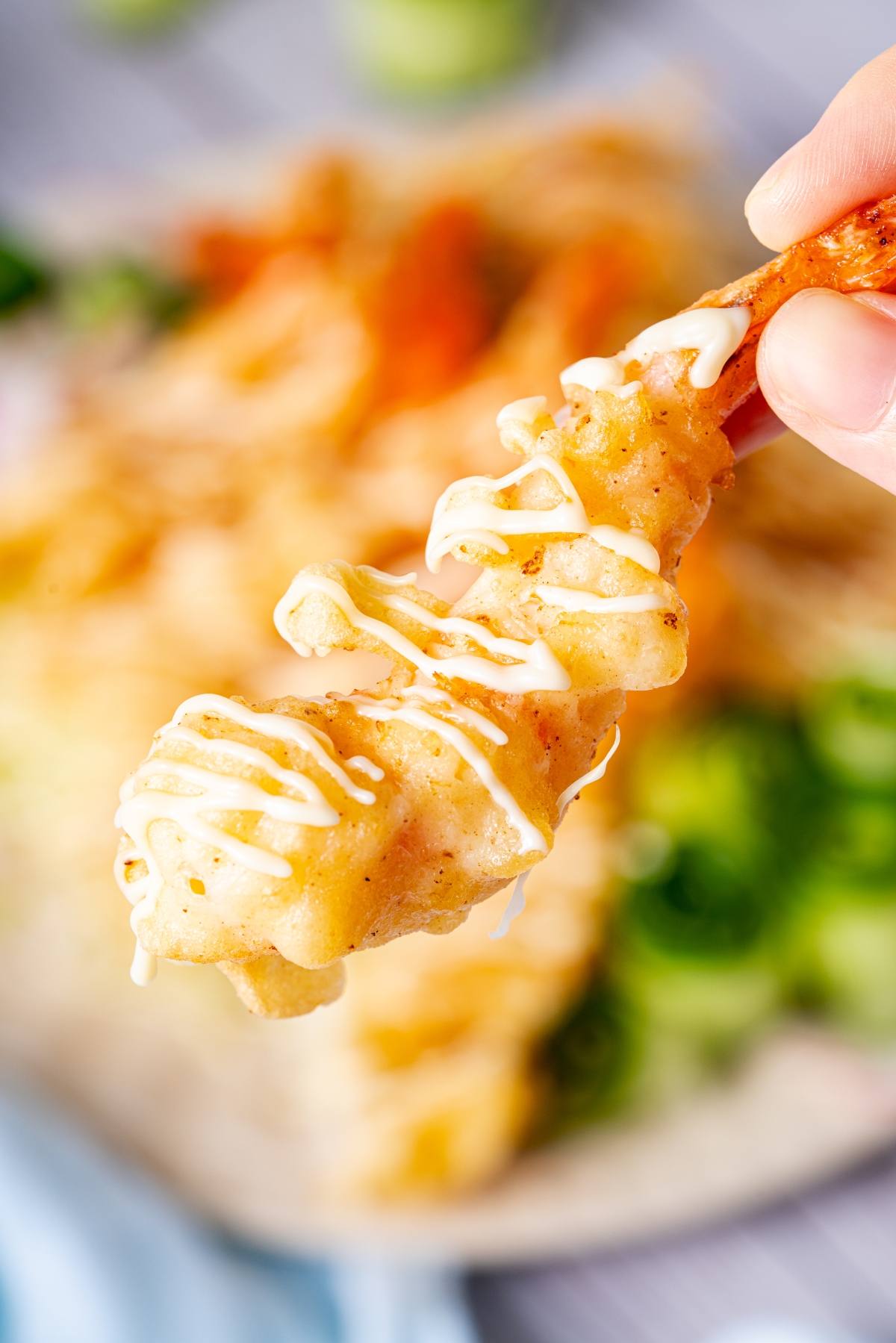 A hand holds a battered shrimp drizzled with creamy white sauce, with blurred food in the background.