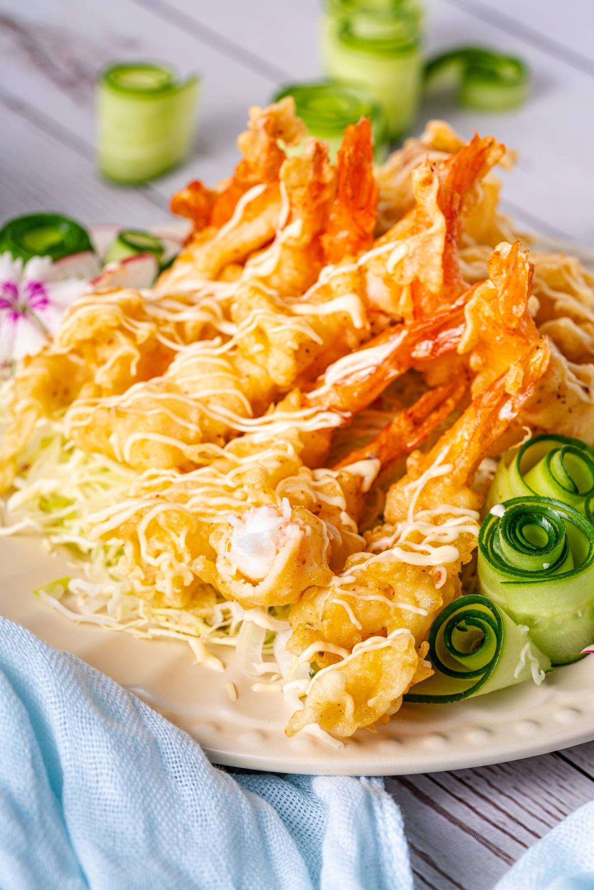 Plate of tempura shrimp drizzled with sauce, served on shredded cabbage with cucumber rolls and flower garnish.
