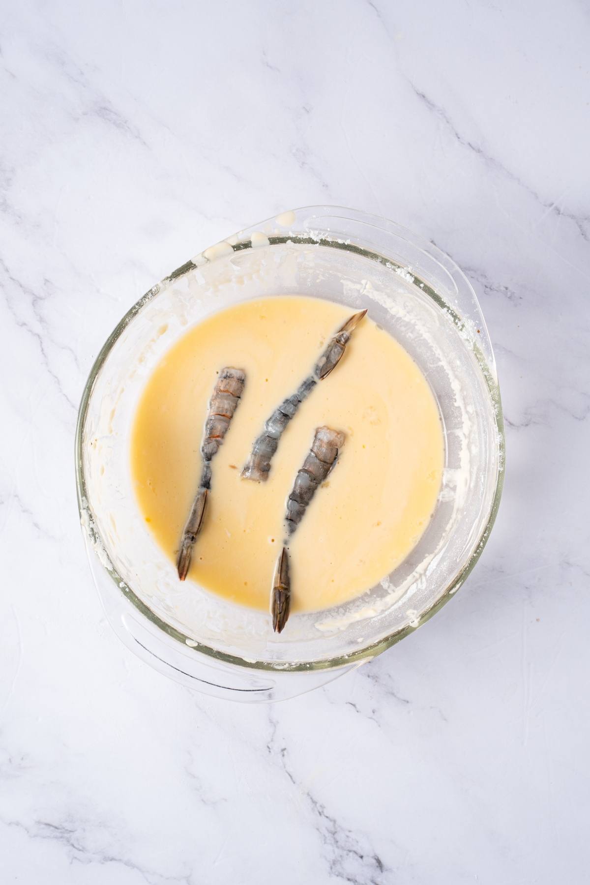 Three raw shrimp in a bowl of batter on a white marble surface.