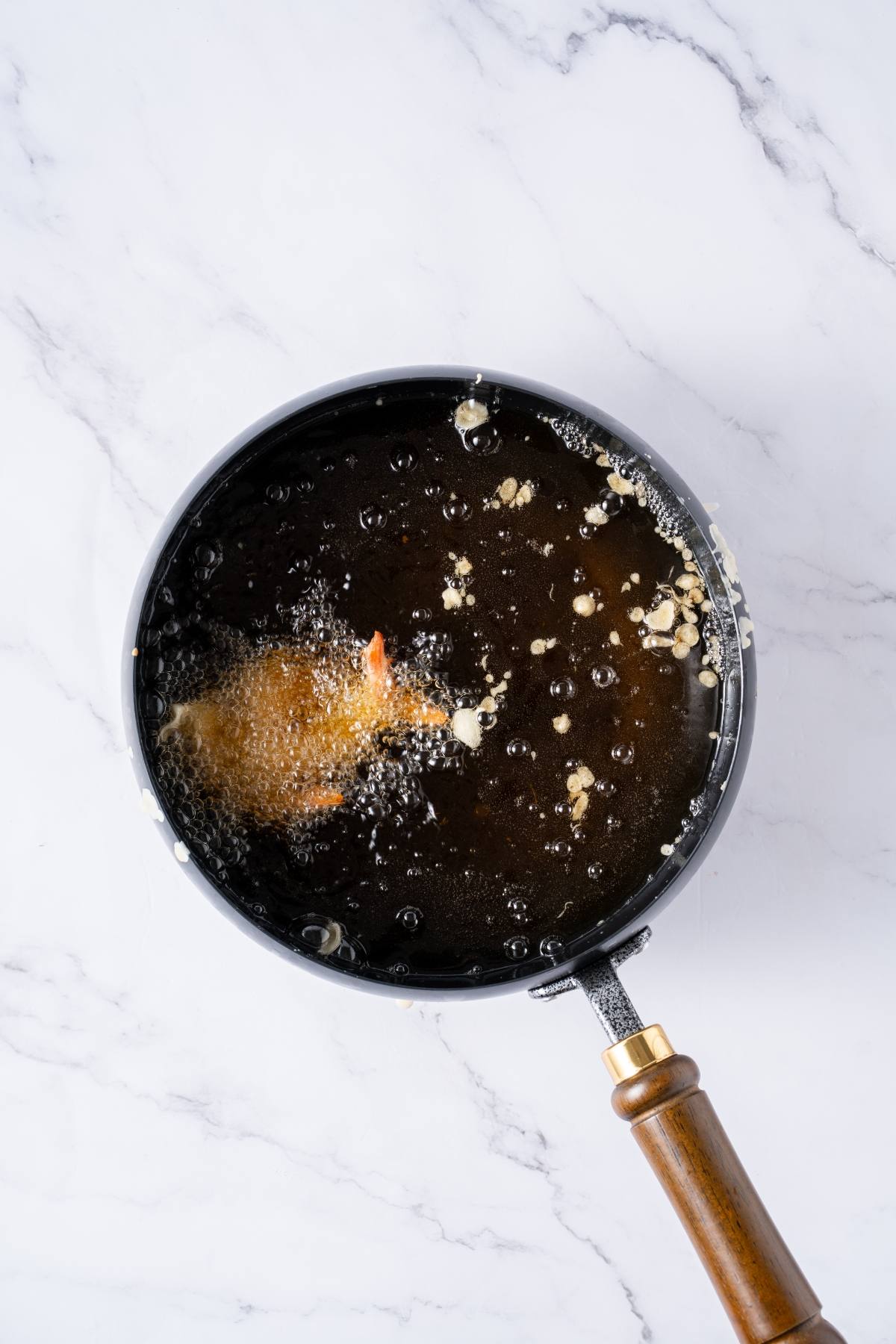 A battered food item frying in hot oil inside a black pan on a white marble surface.