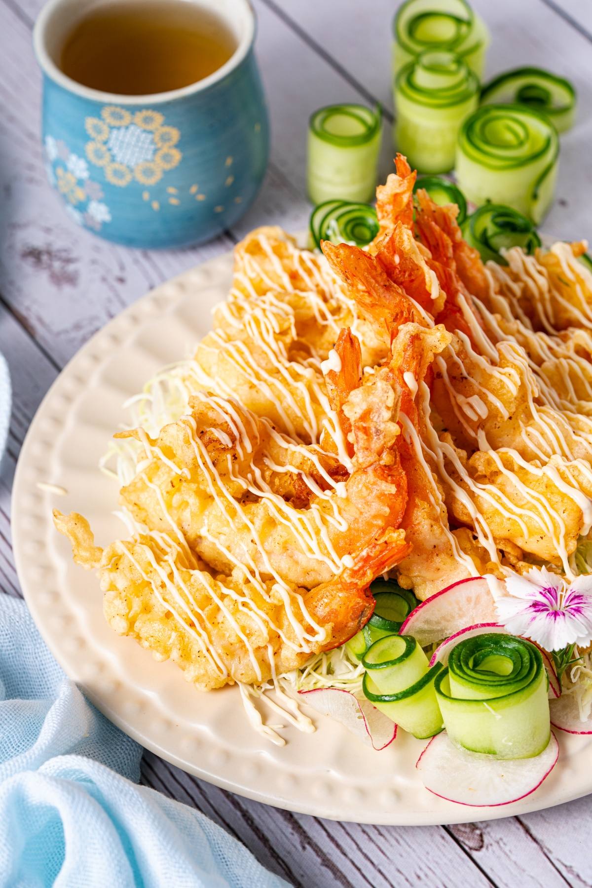 Tempura shrimp with drizzled sauce, served with cucumber ribbons and radish on a white plate.