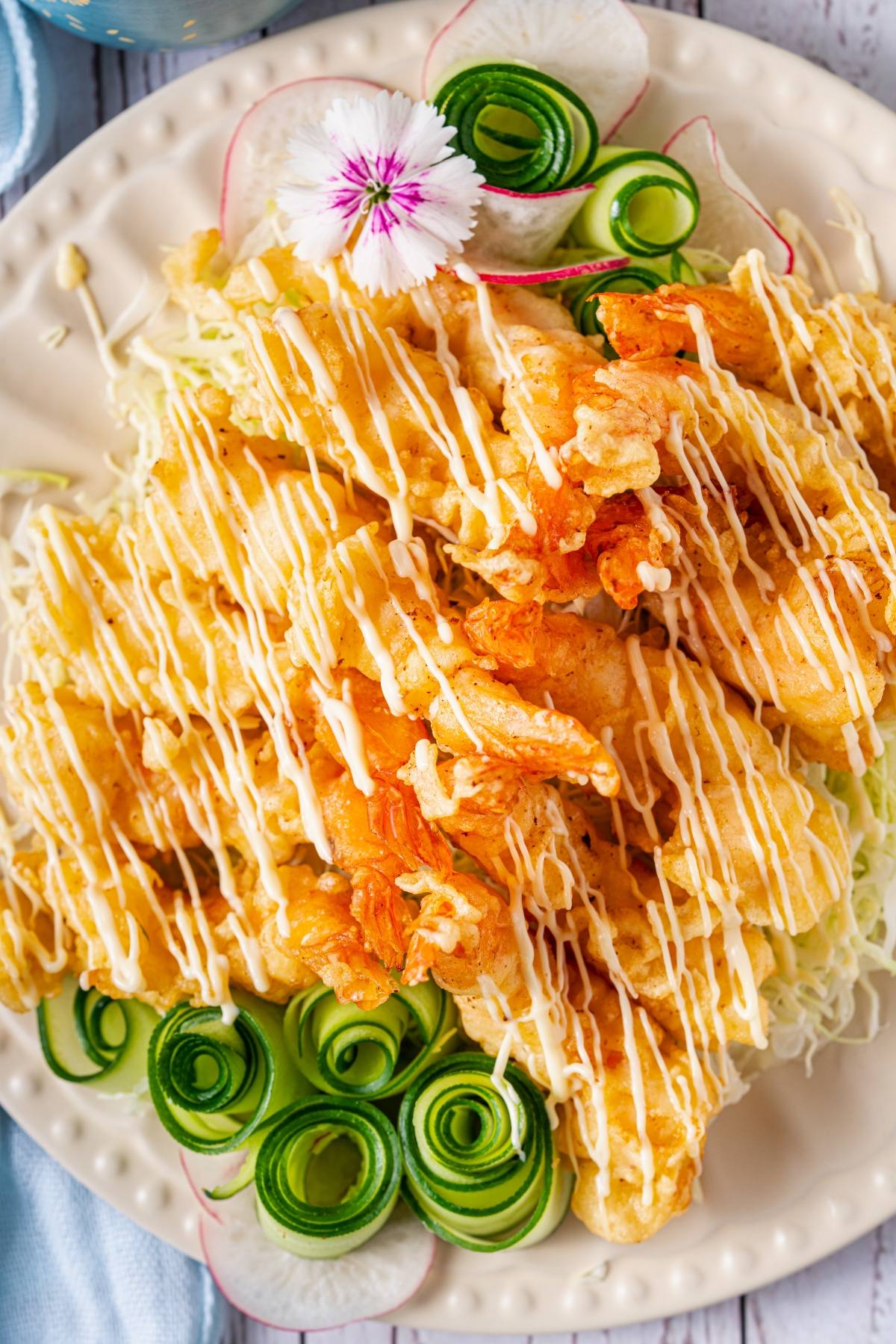 Plate of fried shrimp drizzled with sauce, garnished with cucumber spirals, radish slices, and an edible flower.