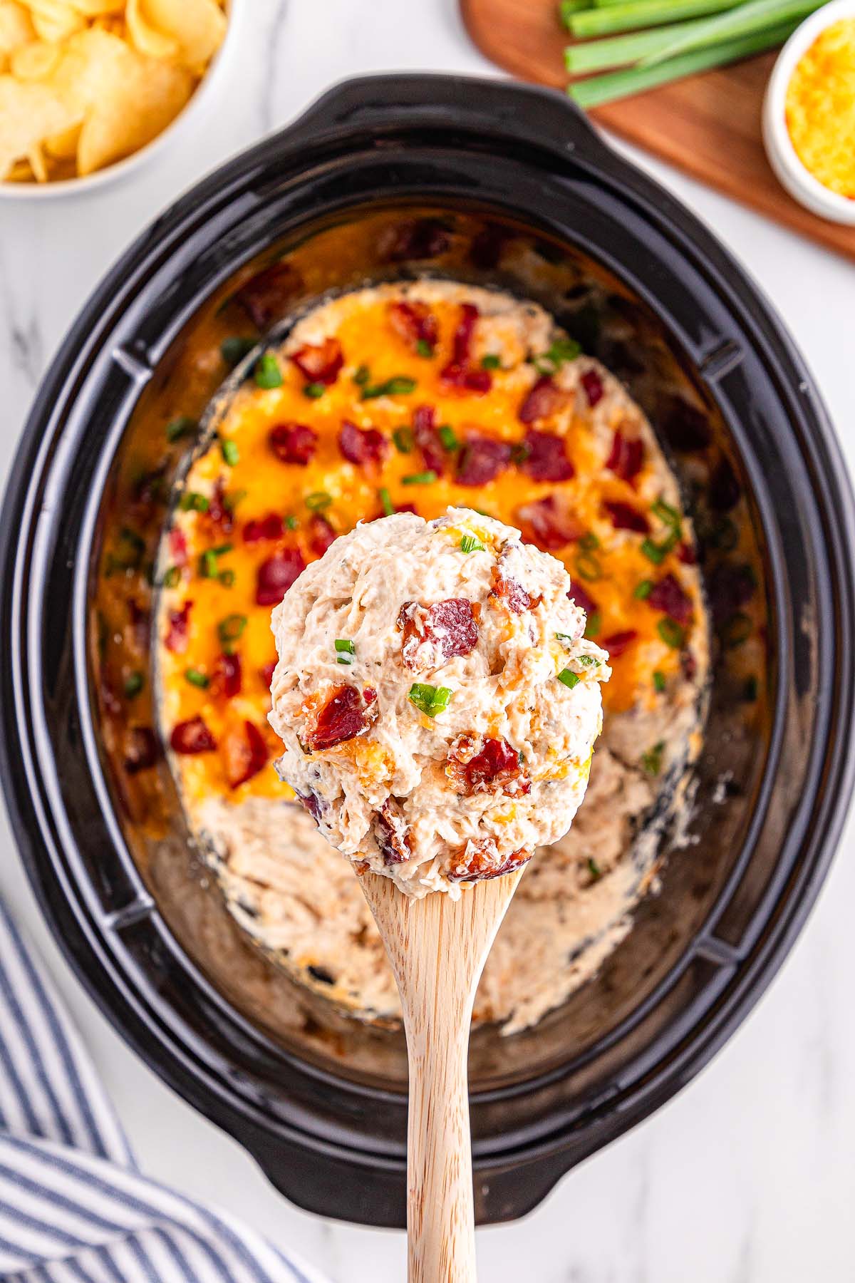 A wooden spoon holds cheesy Slow Cooker Crack Chicken above a slow cooker filled with the same dip, topped with crispy bacon and fresh herbs.