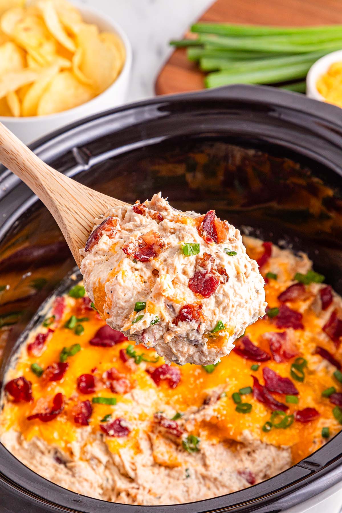 A wooden spoon lifts cheesy Slow Cooker Crack Chicken from a slow cooker, topped with green onions and bacon bits.