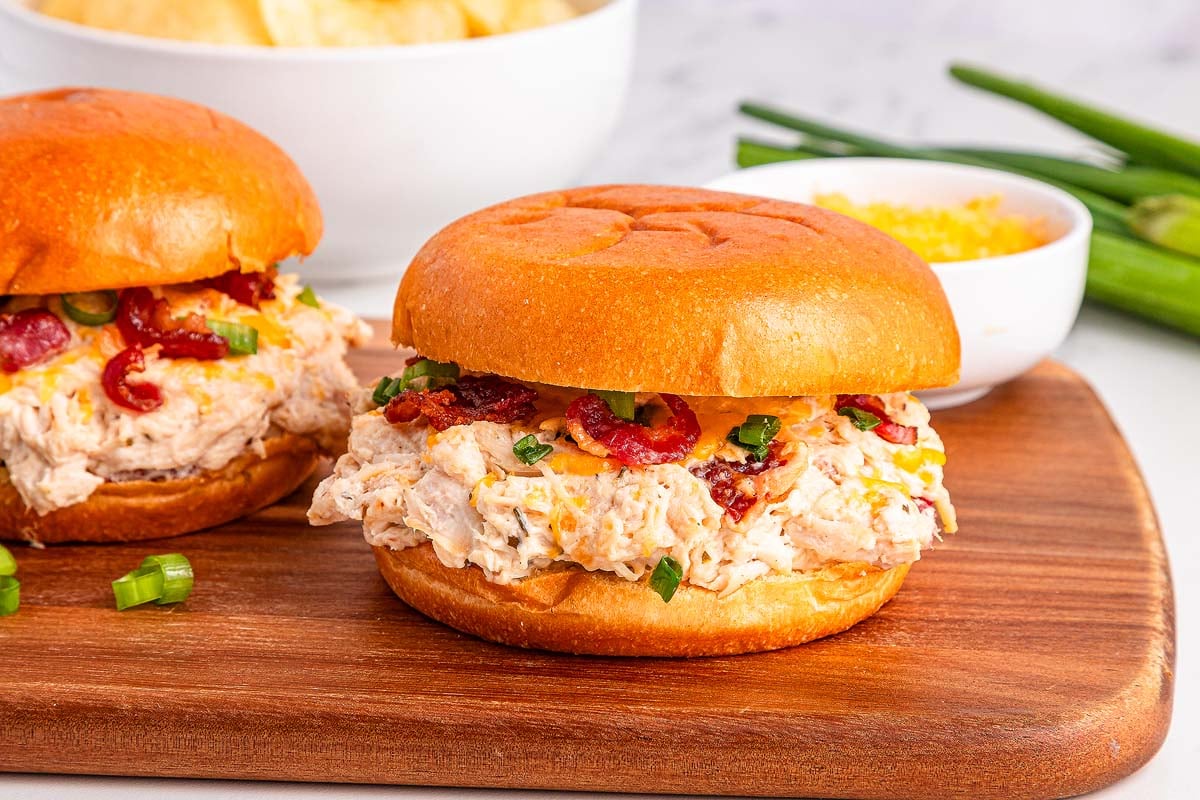 Slow Cooker Crack Chicken on a brioche bun with bacon and green onions, served on a wooden board.