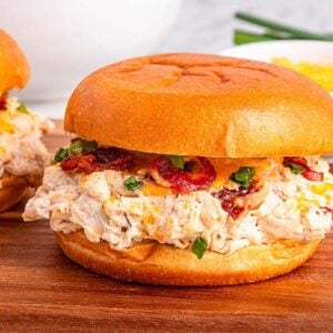 Slow Cooker Crack Chicken on buns with bacon and green onions on a wooden board, with a bowl of cheese in the background.