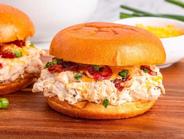 Slow Cooker Crack Chicken on buns with bacon and green onions on a wooden board, with a bowl of cheese in the background.
