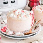 Two mugs of Slow Cooker Candy Cane Hot Chocolate topped with mini marshmallows and peppermint sticks, surrounded by festive holiday decor.