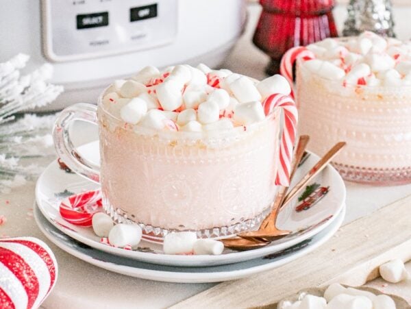 Two mugs of Slow Cooker Candy Cane Hot Chocolate topped with mini marshmallows and peppermint sticks, surrounded by festive holiday decor.