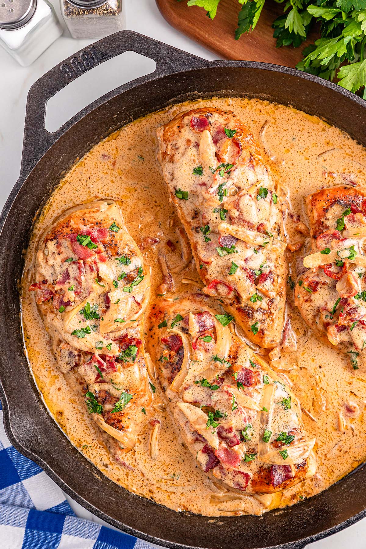 Four creamy Smothered Chicken breasts in a skillet, topped with onions, herbs, and bacon.