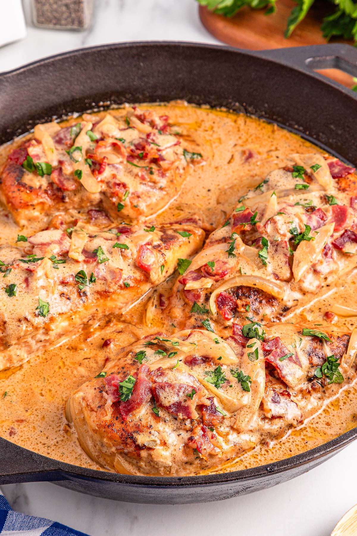 Smothered Chicken breasts simmered in a creamy sauce with onions, herbs, and crispy bacon in a black skillet.