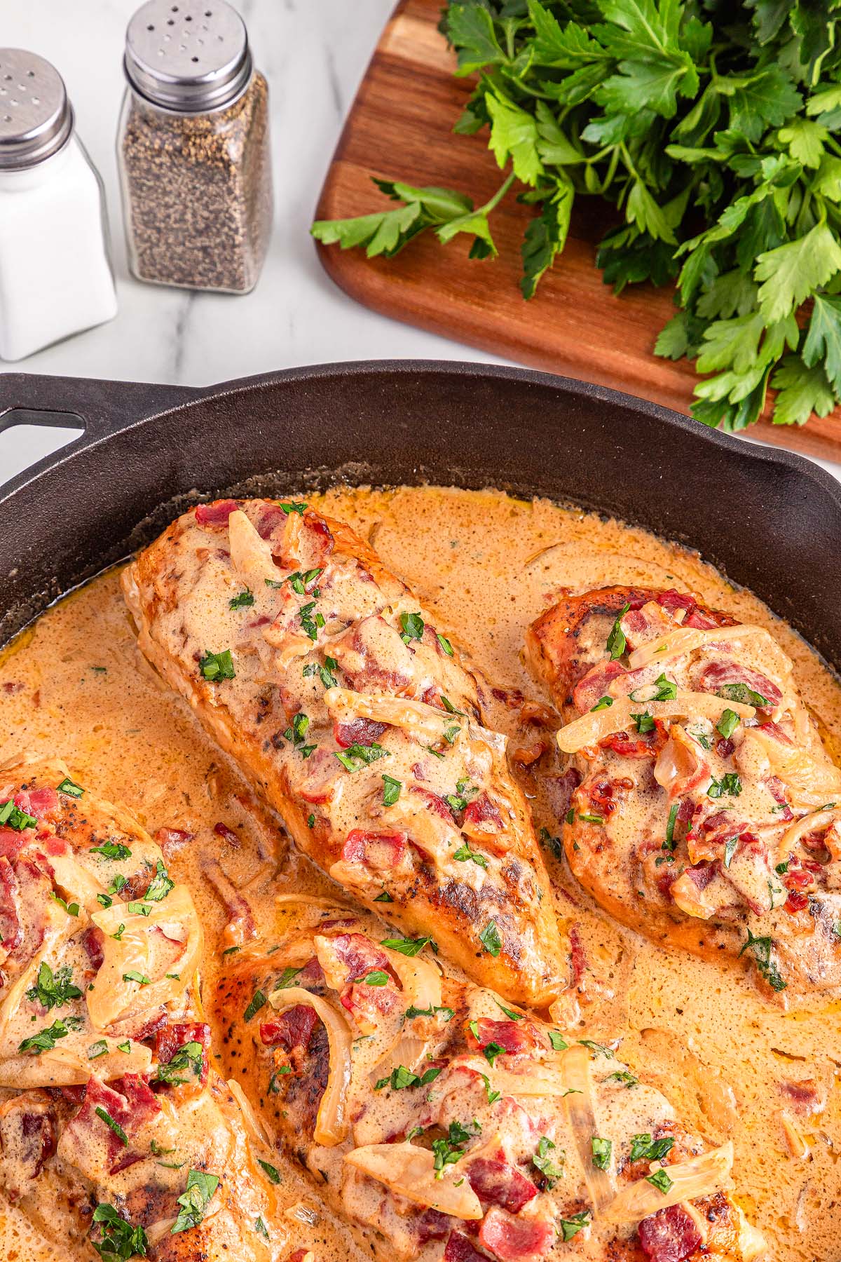 Smothered Chicken breasts in a creamy sauce with herbs, onions, and bacon in a skillet, garnished with chopped parsley.