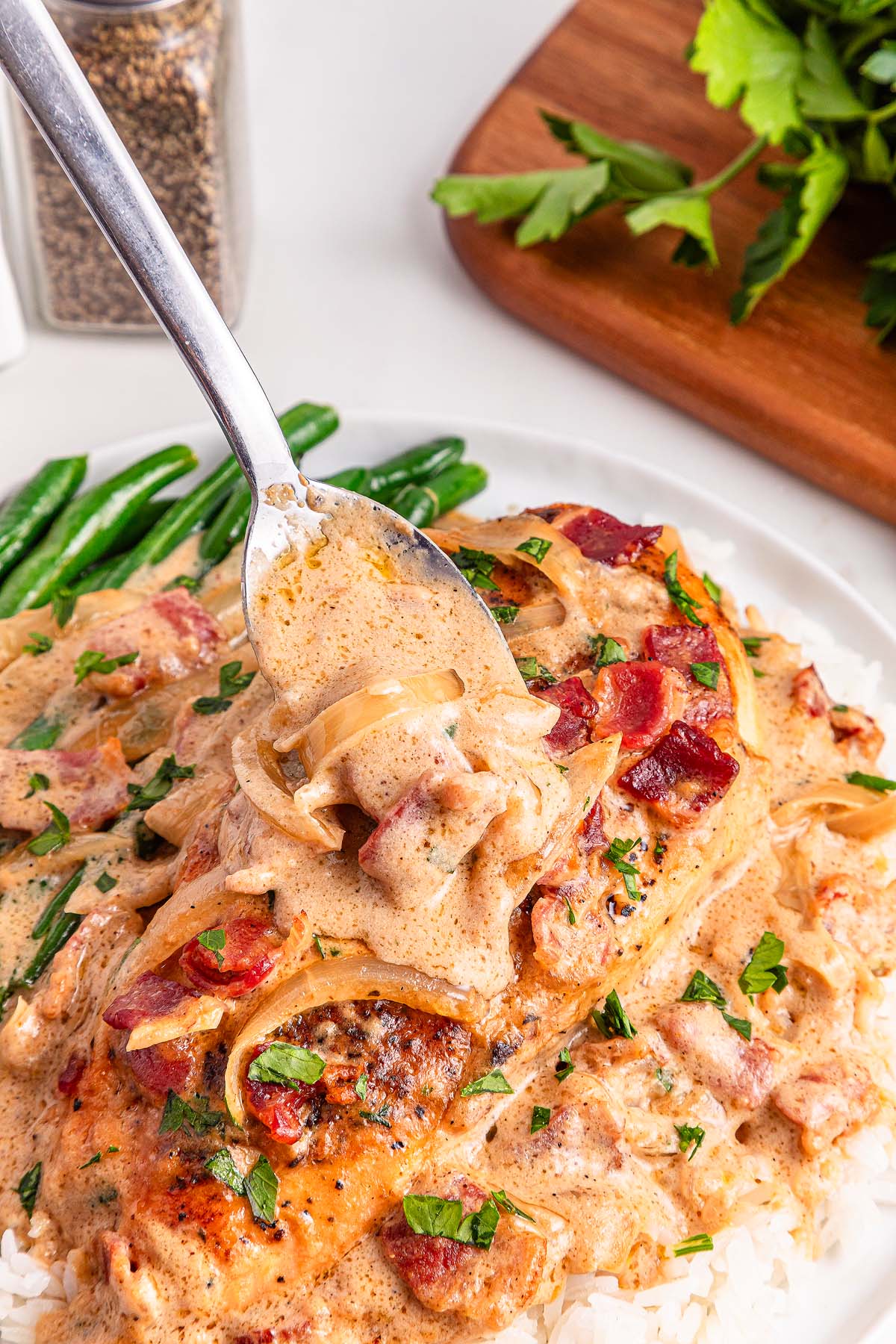 A fork lifts creamy sauce from Smothered Chicken with bacon, onions, and herbs over rice, served alongside green beans.